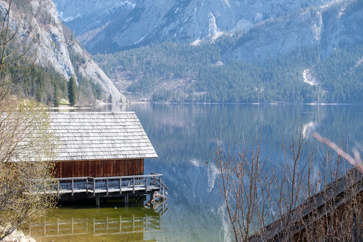 A shingle roofed boathouse on stilts with a wooden deck juts into the clear green water of the Altausseer See, with grey cliffs and forest reflected on the still surface beyond.