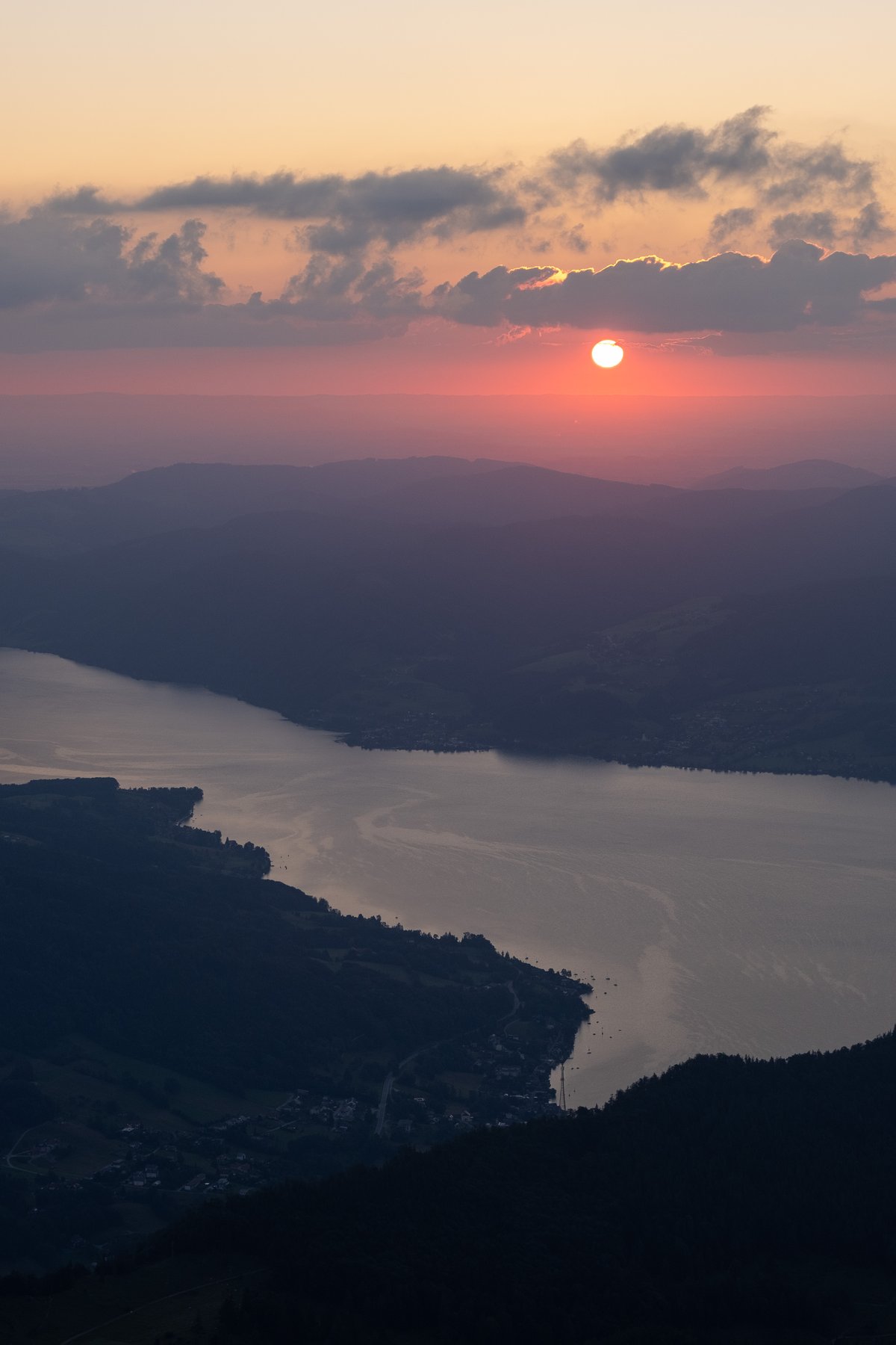 The sun clears a band of cloud above the Attersee at sunrise, seen in telephoto from the Schafberg with the long lake and its shoreline villages far below.