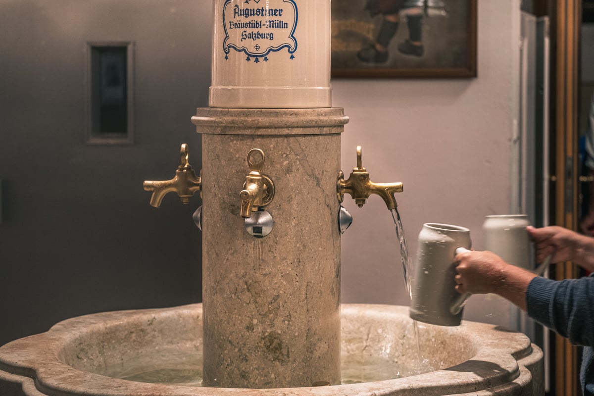 A guest rinses a grey stoneware beer mug under the brass taps of the marble fountain at the Augustiner Braustubl in Mulln, Salzburg, below a display mug lettered in blue with the brewery's name.