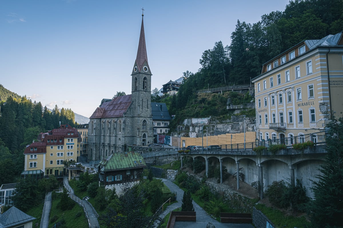 Early morning over Bad Gastein: the stone parish church and its tall spire at the centre, the yellow Kurhotel Mirabell on the right and a concrete road viaduct running below it.