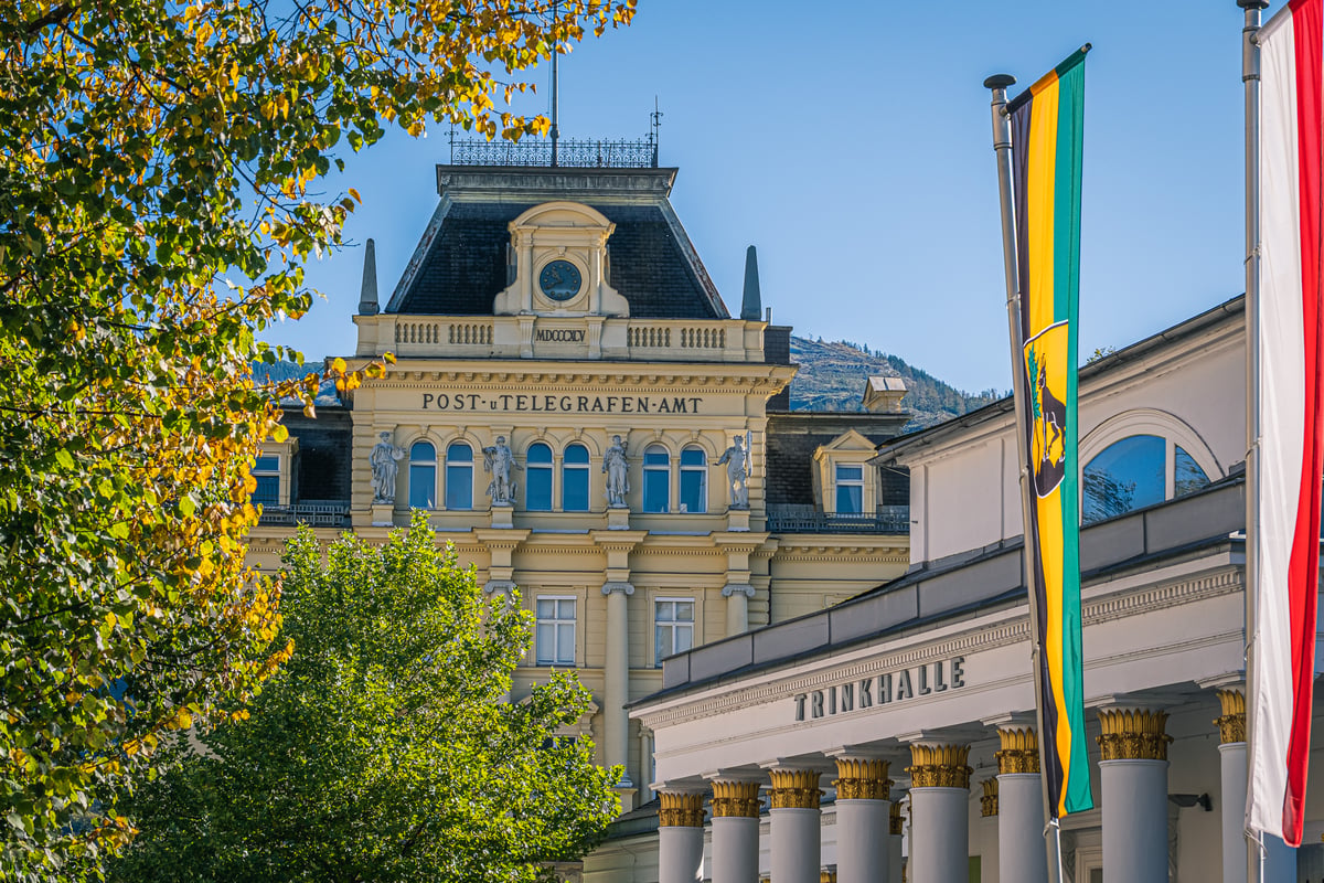 The ochre Post und Telegrafenamt of Bad Ischl, dated MDCCCXCV and topped by a clock gable, rises behind the columned Trinkhalle, framed by autumn foliage under a blue sky.