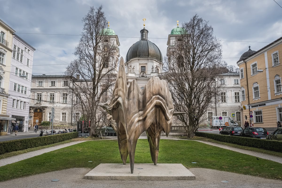 The bronze sculpture Caldera stands on its stone slab on the lawn at Makartplatz in Salzburg, its stacked folds framed by bare late-winter trees with the domed Dreifaltigkeitskirche rising behind.