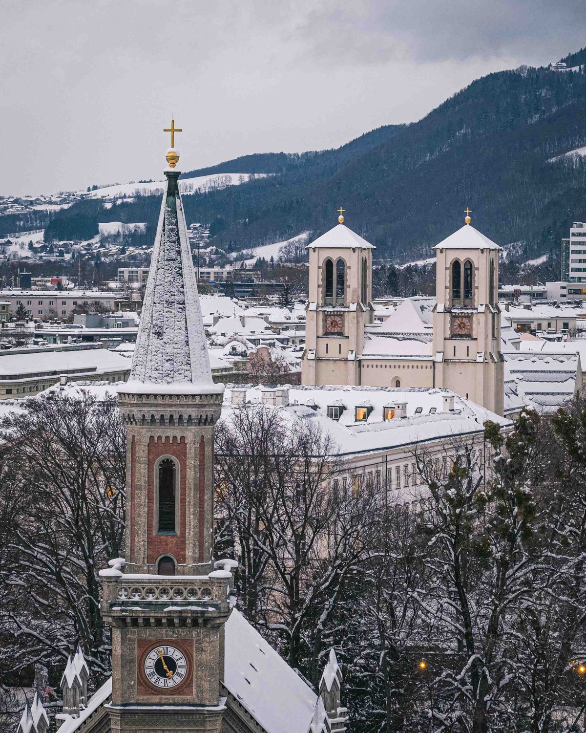 Christuskirche spire with Andrakirche twin towers and snowy Kapuzinerberg hills behind in Salzburg.