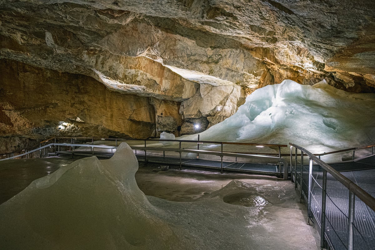 A pale blue dome of ice fills the right side of a wide cave chamber while a railed metal walkway runs across the frozen floor beneath the ochre rock wall.