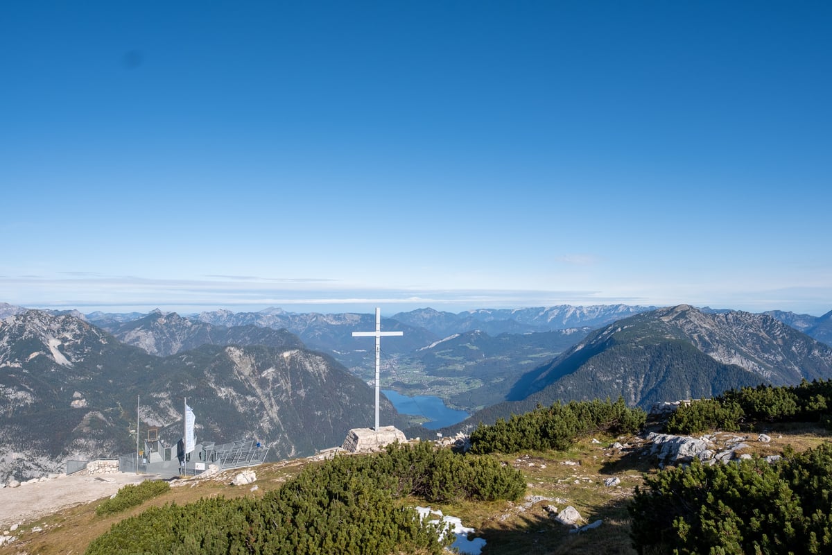 A white metal summit cross stands on the Krippenstein plateau above Obertraun, with the Five Fingers viewing platform on the shoulder to the left and the Hallstättersee lying far below in the valley.