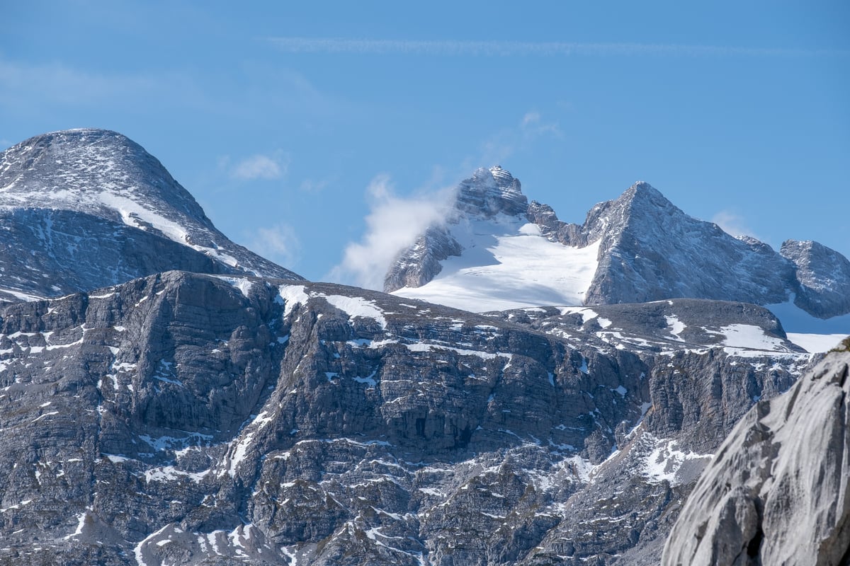 Telephoto view of the Hoher Dachstein summit with cloud streaming off it and the remaining glacier ice spread below, seen across the ridged grey limestone of the plateau.