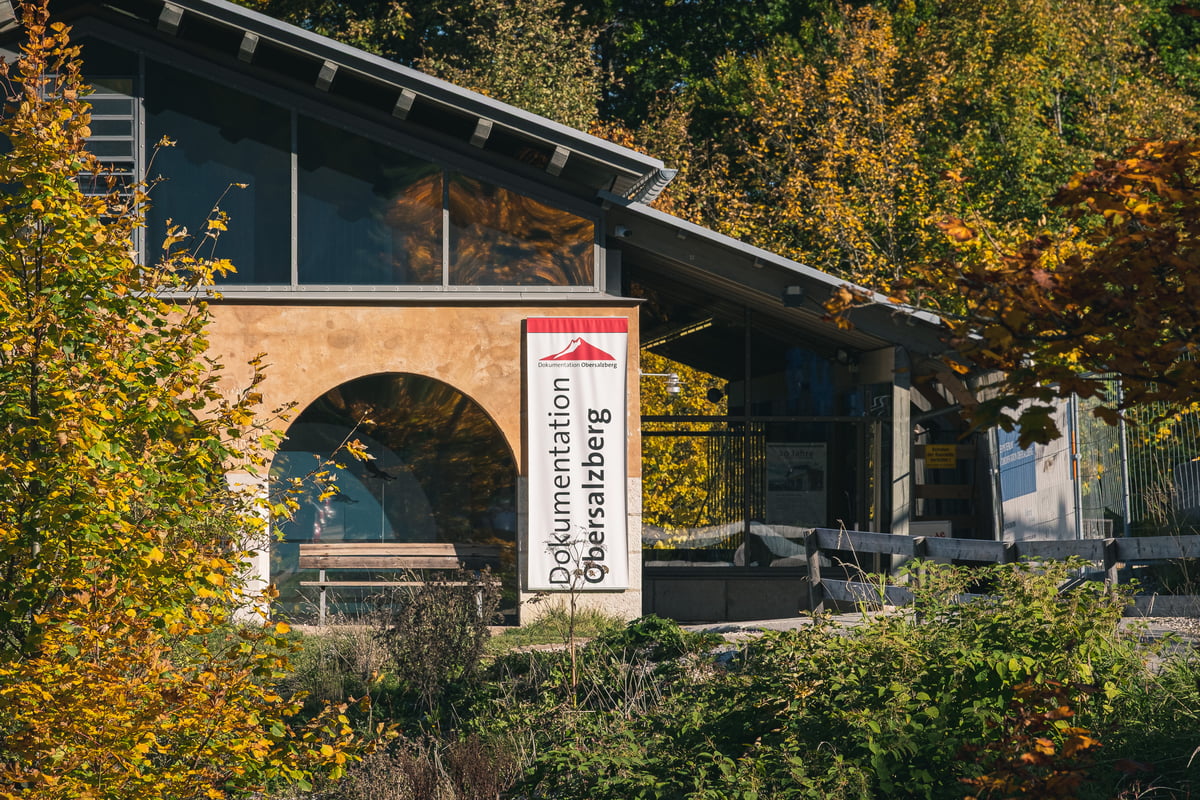 The low modern entrance building of the Dokumentation Obersalzberg, with an arched glazed opening and a tall vertical banner reading Dokumentation Obersalzberg, surrounded by yellowing autumn trees.