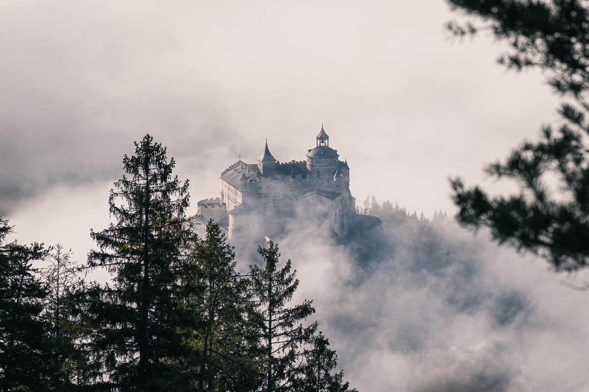 Festung Hohenwerfen stands on its wooded rock with its onion-domed bell tower and battlements rising out of a thick bank of morning fog, seen past dark spruce tops.