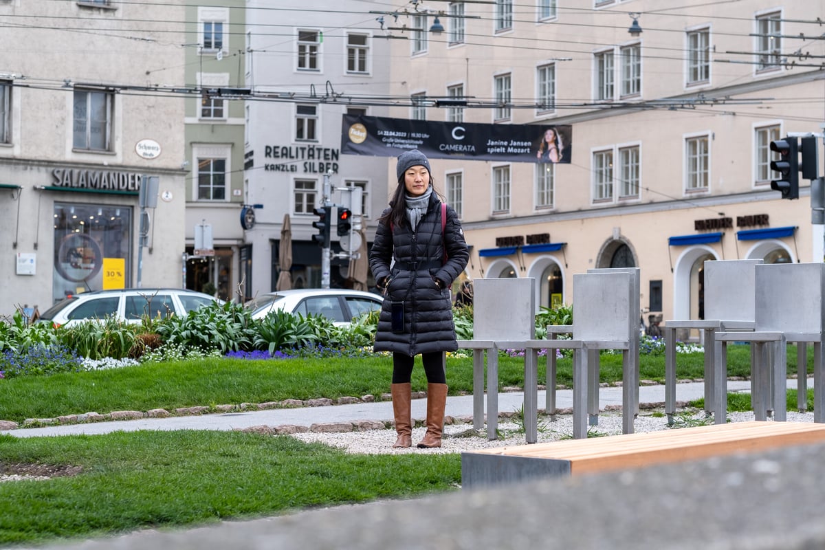 A Free Walking Tour Salzburg guide in a long black coat and brown boots stands on a lawn among the tall steel chairs of the Spirit of Mozart installation, with shopfronts and trolleybus wires behind.