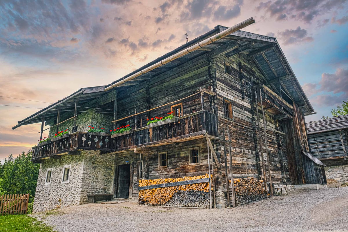 Large wooden alpine farmhouse with balcony and firewood stacks under evening sky at the open-air museum near Salzburg.