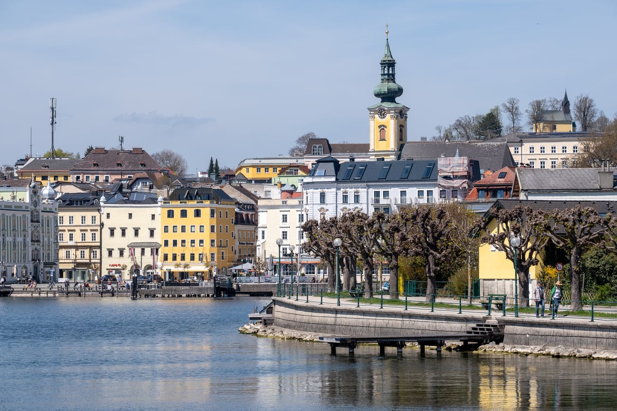 The Gmunden lakefront in spring, with the pastel Rathausplatz houses and the yellow-towered parish church on the left and the Esplanade promenade curving along the right behind pollarded trees, all seen from the Traunsee.