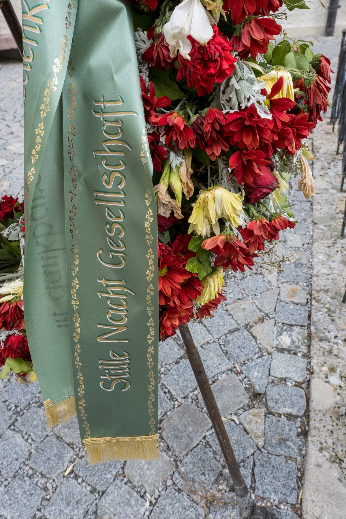 A memorial wreath of red and yellow flowers with a green sash lettered Stille Nacht Gesellschaft in gold, standing on cobblestones in Hallein.