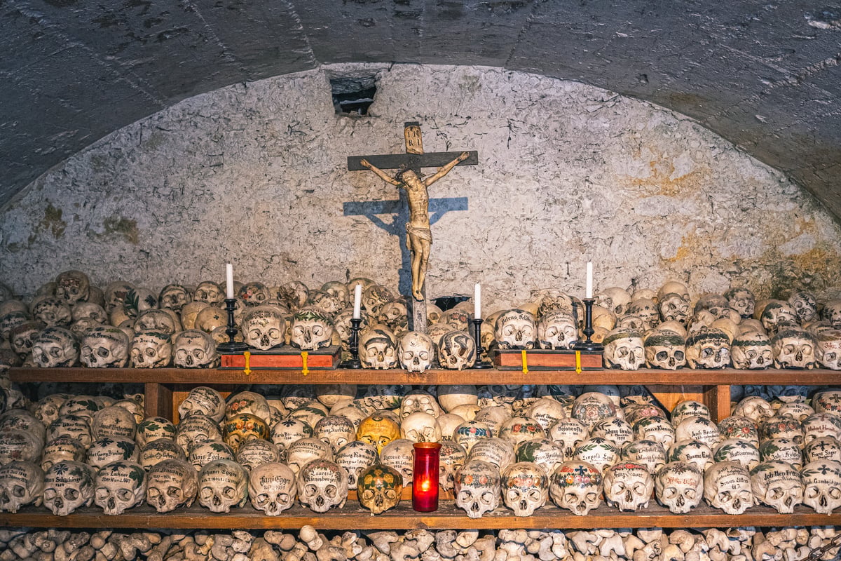 The vaulted interior of the Hallstatt charnel house, with a carved crucifix on the whitewashed back wall, two long shelves of painted skulls, standing candles and a red votive lamp, and packed bones along the floor.