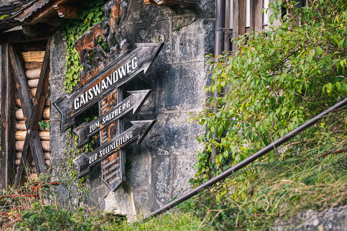 Three weathered wooden arrow signposts reading Gaiswandweg, Zum Salzberg and Zur Solenleitung are fixed to a stone wall in Hallstatt beneath a carved chamois, with a timber woodshed to the left.