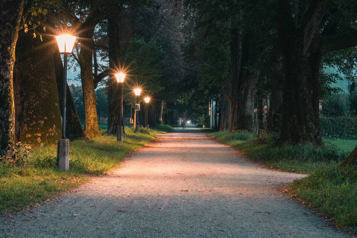 Tree-lined Hellbrunner Allee gravel path with glowing vintage lamps at dusk in Salzburg