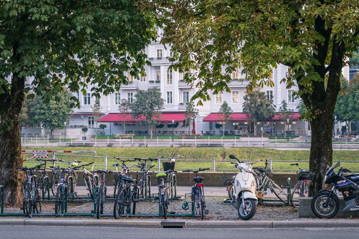 Bikes and scooters parked below chestnut trees opposite Hotel Sacher Salzburg with red awnings in Salzburg
