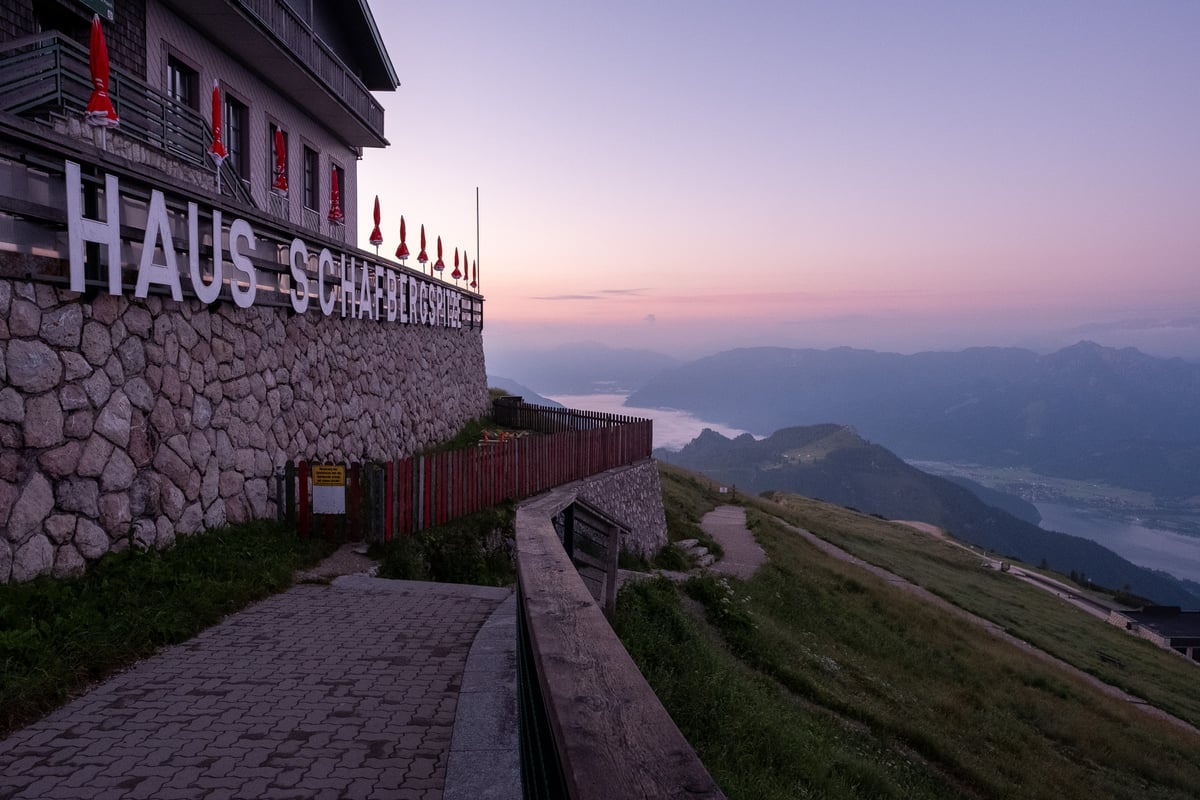 Tall white letters spelling Haus Schafbergspitze run along the stone base of the summit hotel on the Schafberg, with furled red umbrellas above the terrace and a pink dawn sky over the Salzkammergut.