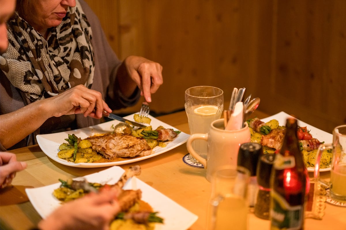 A woman cuts into her pork chop at a pine-panelled restaurant table laid with a lemon-topped beer glass, a ceramic mug of cutlery, pepper mills, bottles and a red candle.