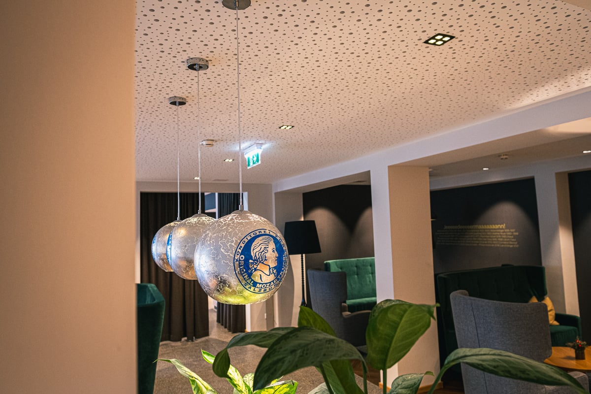 At JUFA Hotel Salzburg, three spherical pendant lamps printed with the blue Original Salzburger Mozartkugel wrapper hang over a hotel lobby, a dark panel headed Jeeeeedeeeermaaaaaann on the wall beyond.
