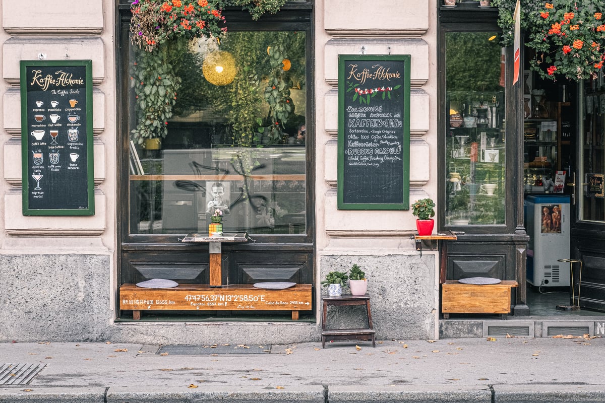 The Kaffee Alchemie storefront in Salzburg with two green-framed chalkboards, a hand-drawn drinks menu on one and a bean list on the other, above window benches and potted plants on the pavement.