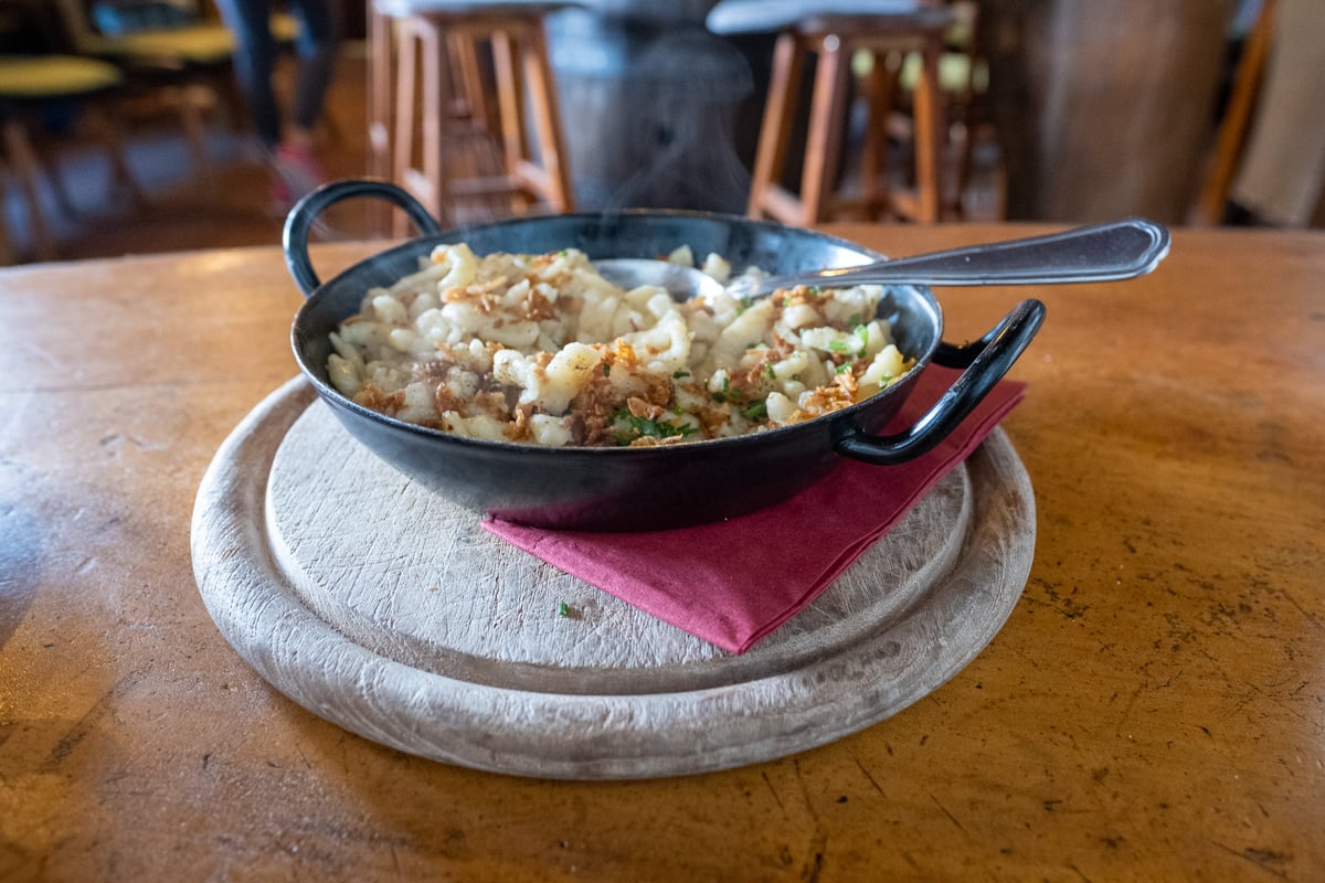 A steaming black iron pan of Kasnocken topped with crisp fried onions and chives sits on a pale wooden board, with the bar stools of a mountain hut behind.