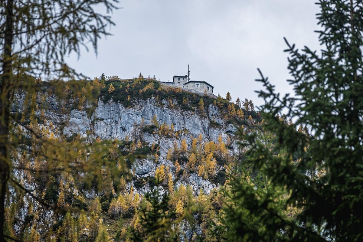 The grey stone Kehlsteinhaus with its aerial mast sits on the very edge of a pale limestone cliff, seen from far below through golden larches and dark spruce under an overcast sky.