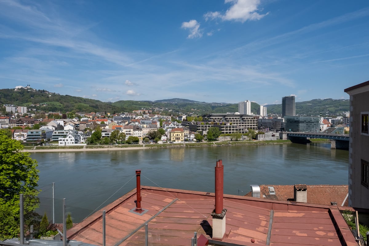 View from the castle hill in Linz over a red tin roof to the Danube, with the Urfahr bank opposite, the Poestlingberg church on the ridge at the left and the Nibelungen bridge at the right.