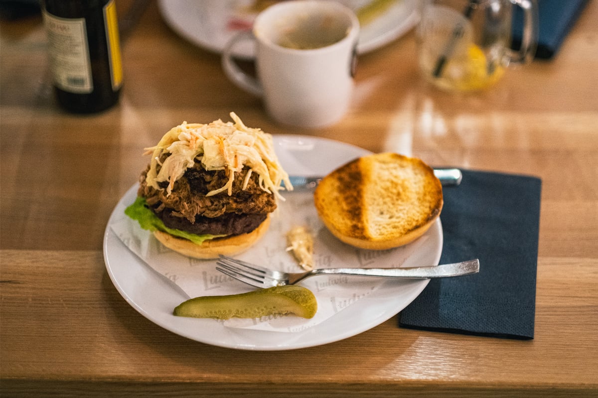 An open pulled pork burger piled with coleslaw sits on a Ludwig-branded paper liner beside a toasted bun lid and a pickle spear.