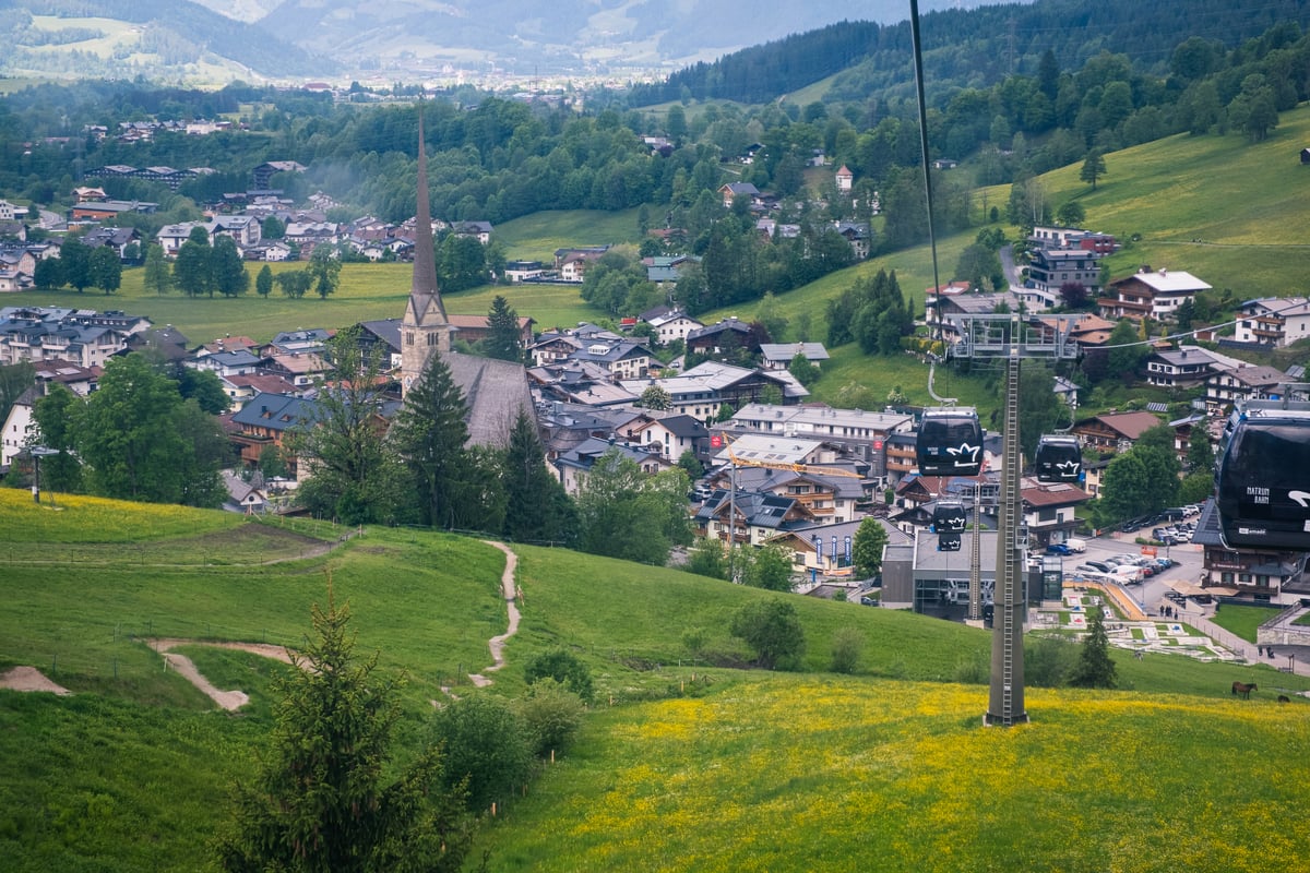 Maria Alm seen from the Natrun hillside in early summer, its tall church spire rising from the village while black Natrunbahn gondolas cross a buttercup meadow in the foreground.