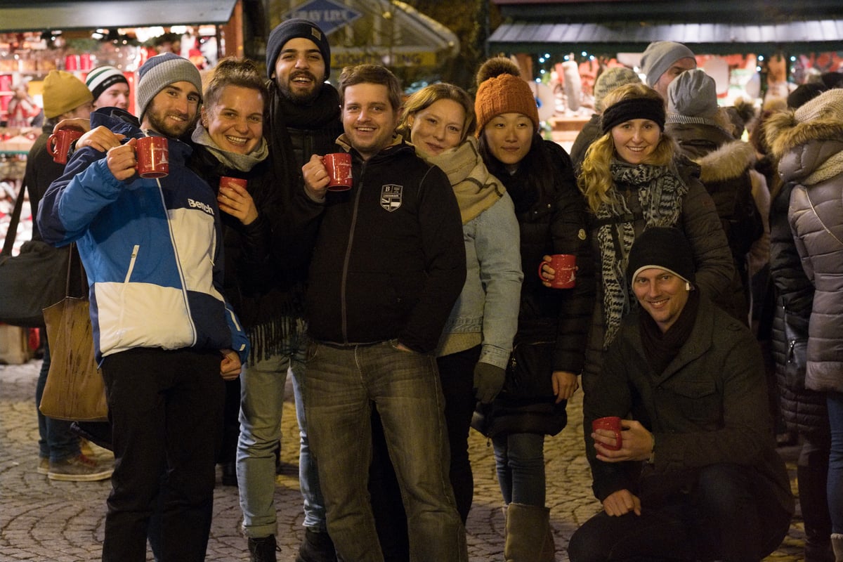 Eight friends in winter hats and coats crowd together for the camera, raising red Gluehwein mugs in front of the lit stalls of the Christmas market at night.