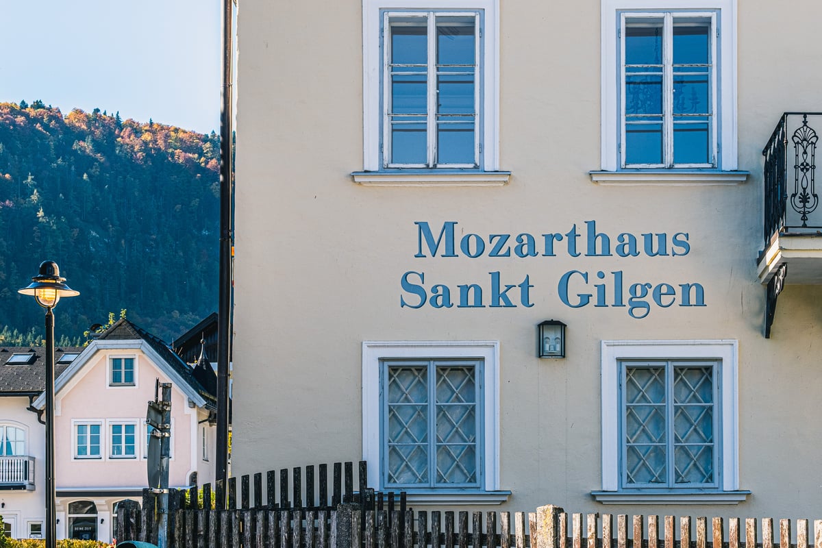 The cream facade of the Mozarthaus in St. Gilgen, its name painted in blue letters between shuttered windows, with a wooded hillside behind a picket fence.