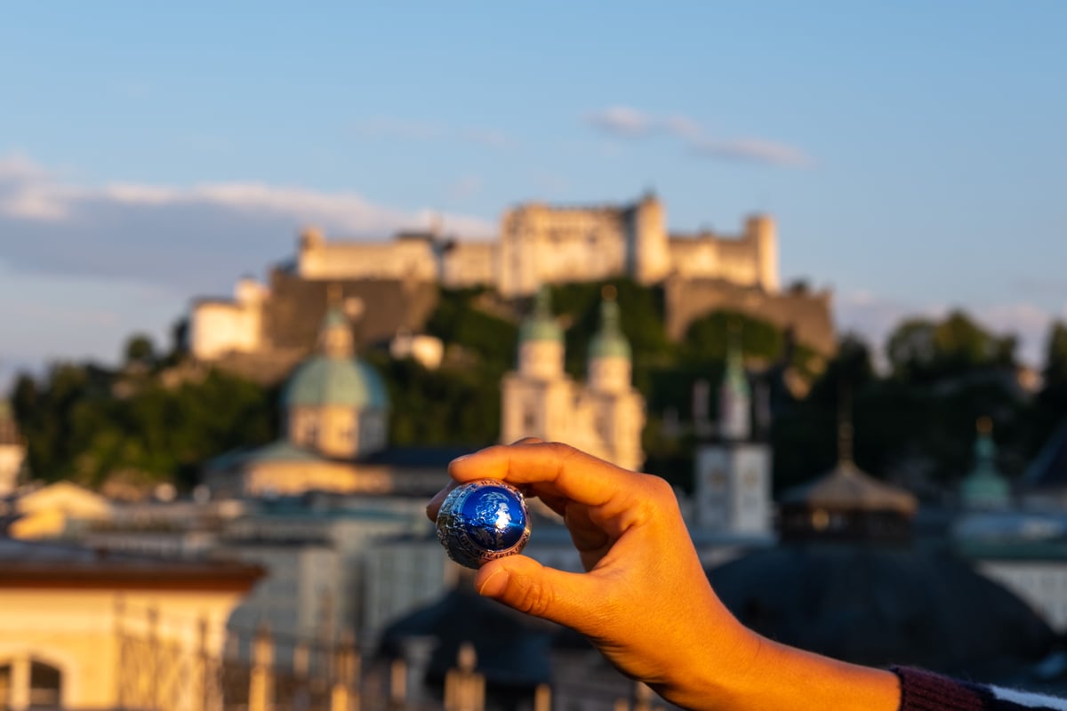 A hand holds a blue-foil Mozartkugel up between finger and thumb against the sunlit Hohensalzburg Fortress and the Salzburg Altstadt skyline.