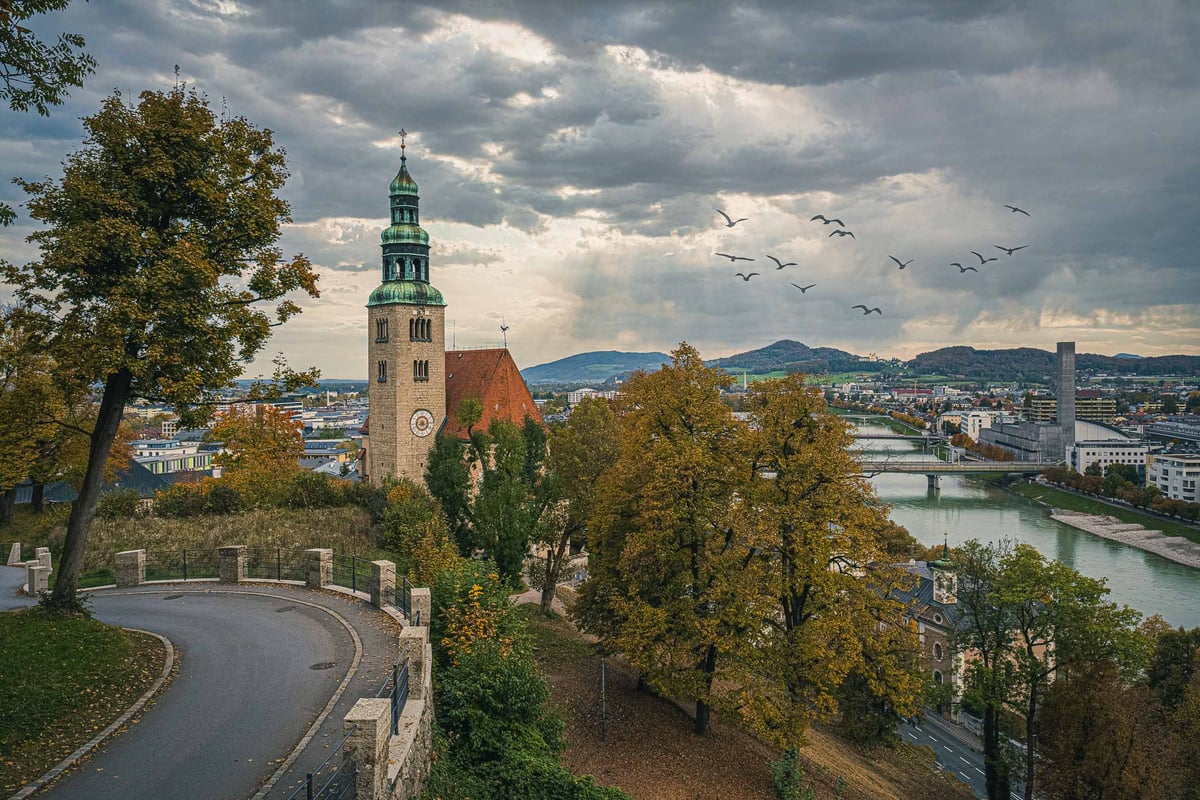 Flock of birds flies past Muelln Church tower above Salzach River with autumn trees in Salzburg.