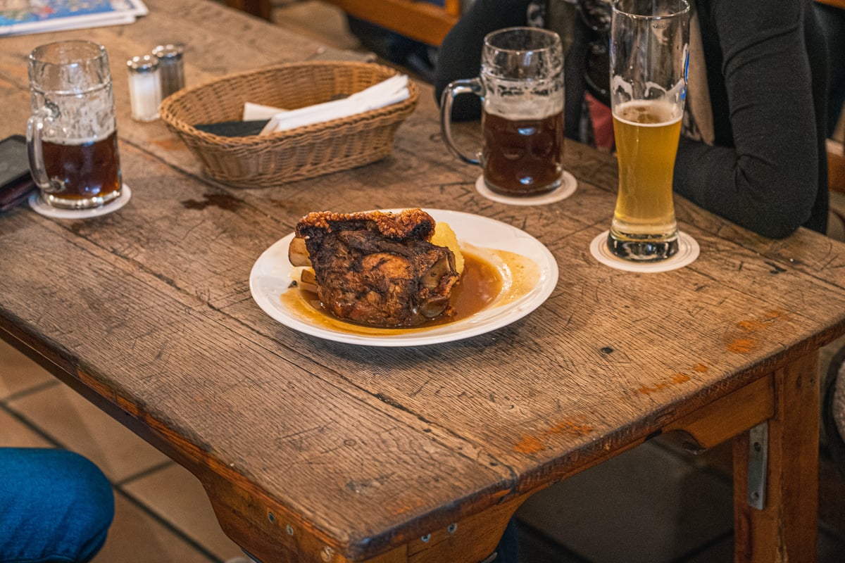 A roast pork knuckle with crackling sits in gravy beside a dumpling on a white plate, on a deeply scratched wooden beer-hall table flanked by glass steins of dark beer and a tall wheat beer.