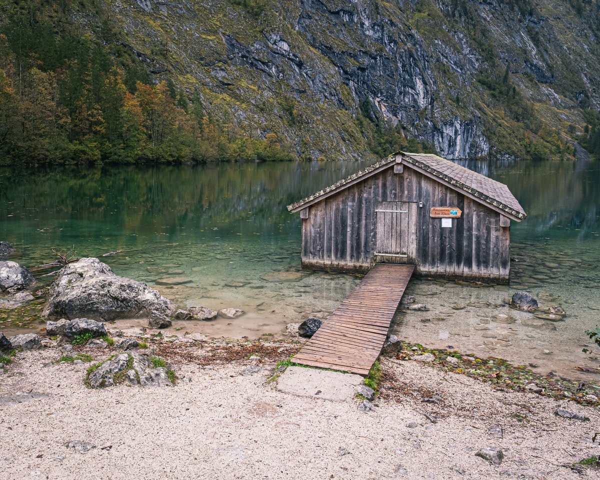 The Obersee boathouse seen across clear turquoise shallows from the pale gravel shore, with autumn woods and grey rock rising behind the lake.