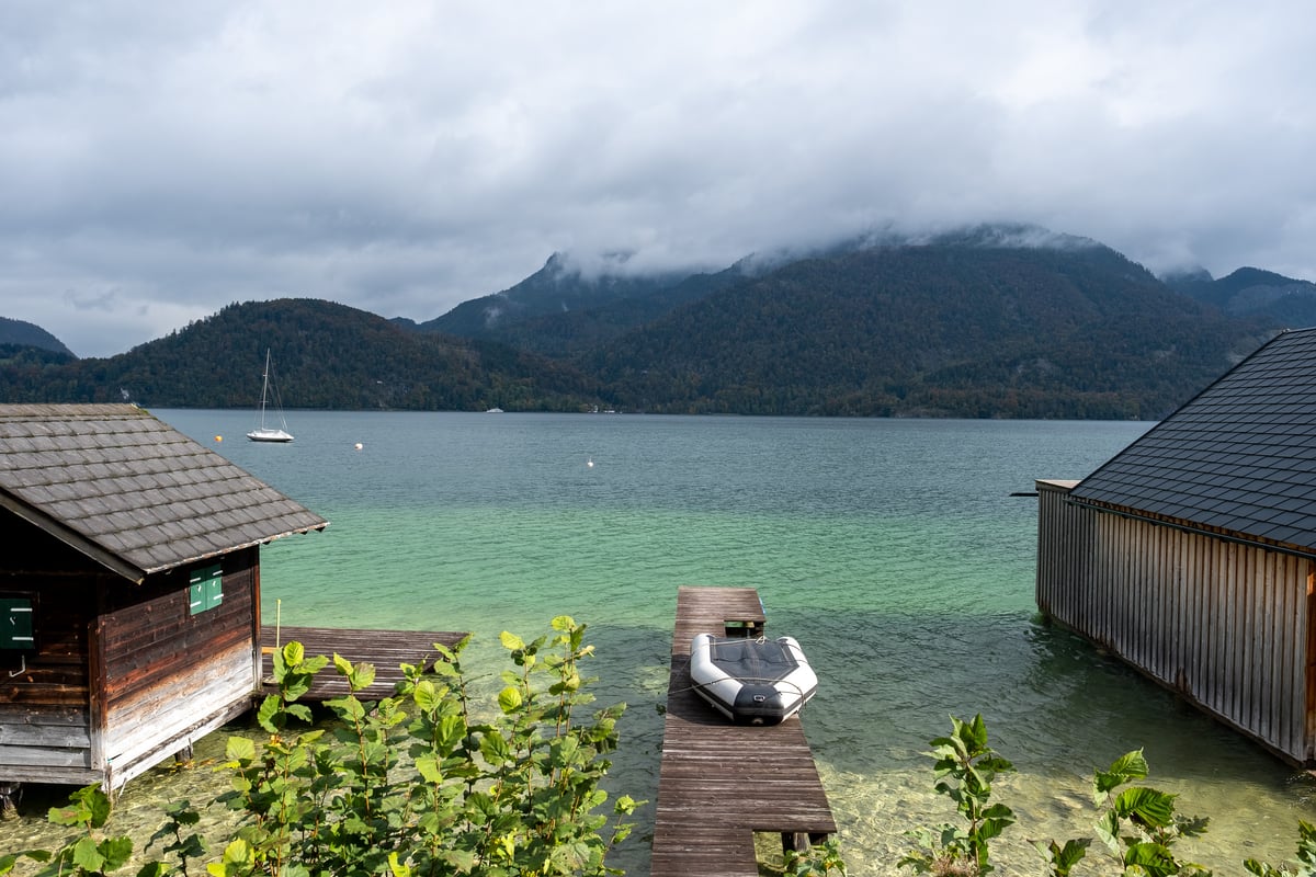 Two timber boathouses flank a plank jetty with a moored dinghy on a green alpine lake, a yacht at anchor beyond and cloud lying low on the wooded ridges.