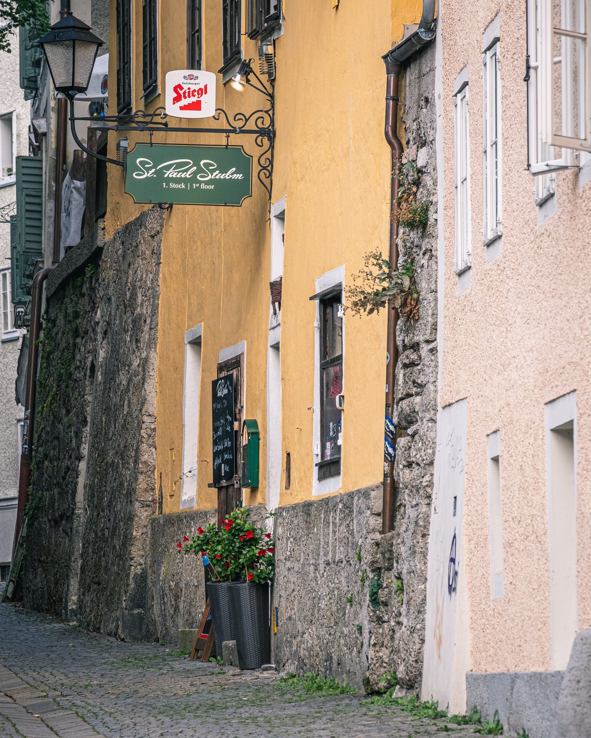 The green St. Paul Stubm sign and a Stiegl plaque on the ochre wall of Pauli Stubm, seen up a narrow cobbled lane past a stone retaining wall and a box of red geraniums in Salzburg.