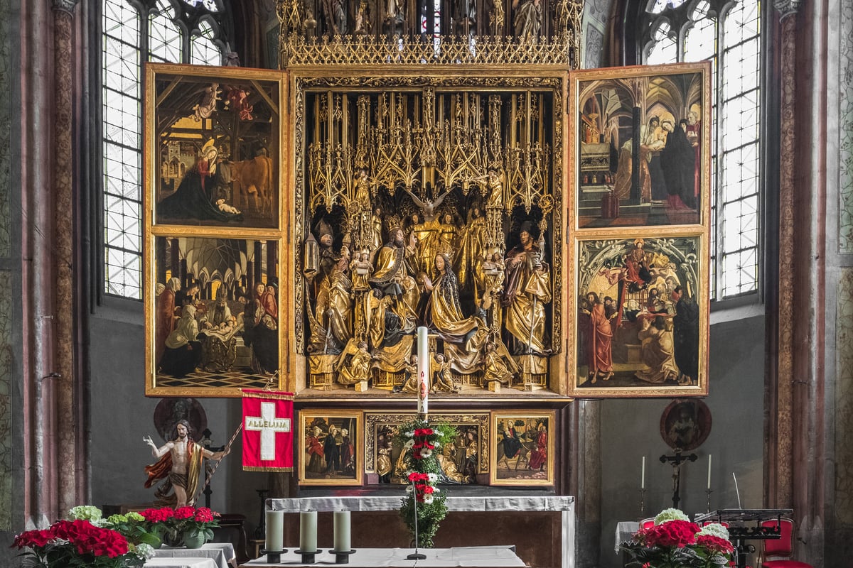The Michael Pacher winged altarpiece in the church of St. Wolfgang stands fully open, its gilded Coronation of the Virgin shrine flanked by painted panels, with a red Alleluja banner, a paschal candle and a Risen Christ figure in front.