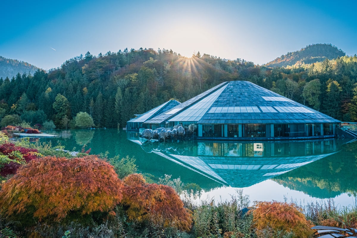 The conical glass and slate buildings of the Red Bull headquarters at Fuschl am See mirrored in a still turquoise lake, with the sun breaking over the forested ridge behind and red maples in the foreground.