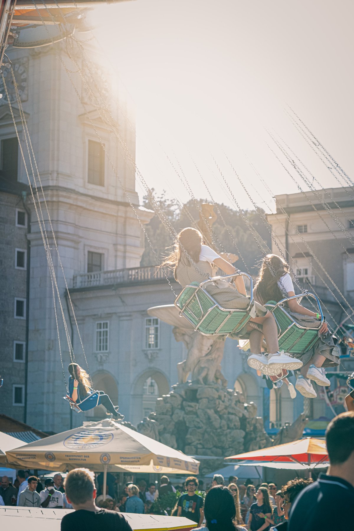 Two young women in dirndl swing out on the chain carousel into low backlit sun, with the Residenzbrunnen and a tower catching the flare behind them.