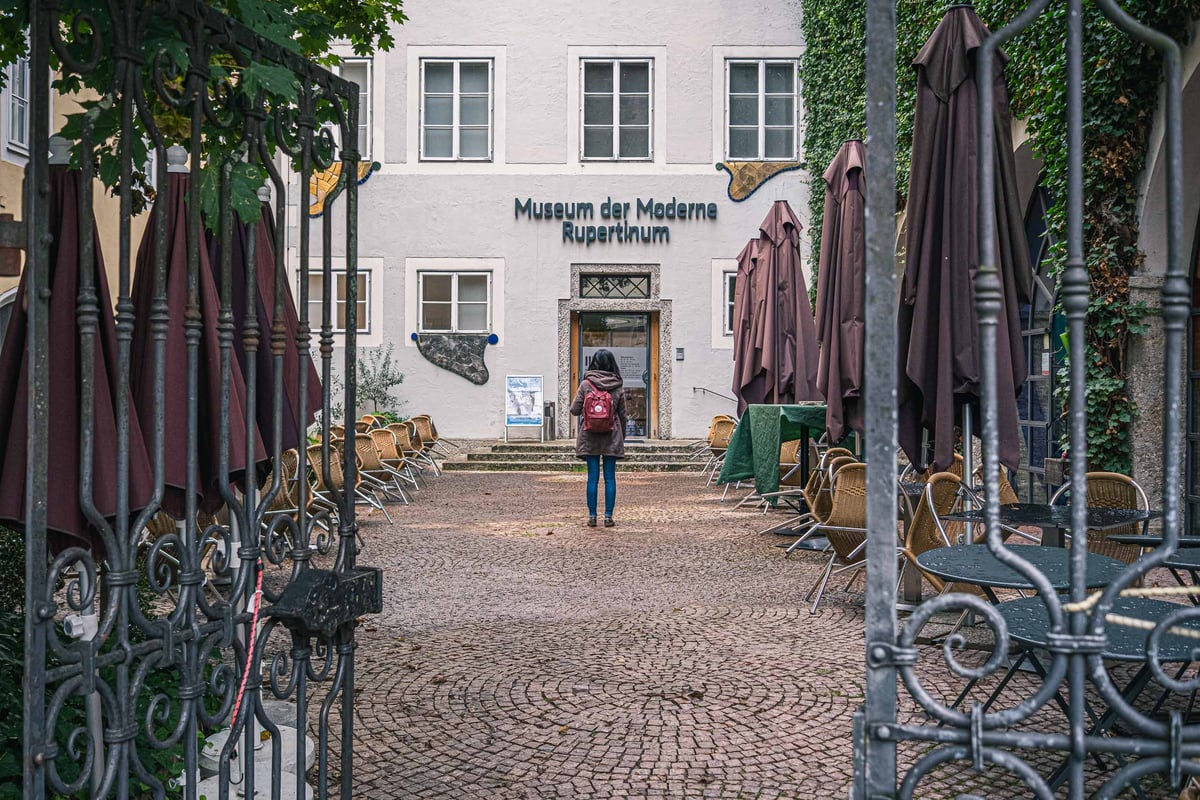 View through an iron gate toward the Museum der Moderne Rupertinum entrance with cafe chairs in Salzburg old town.