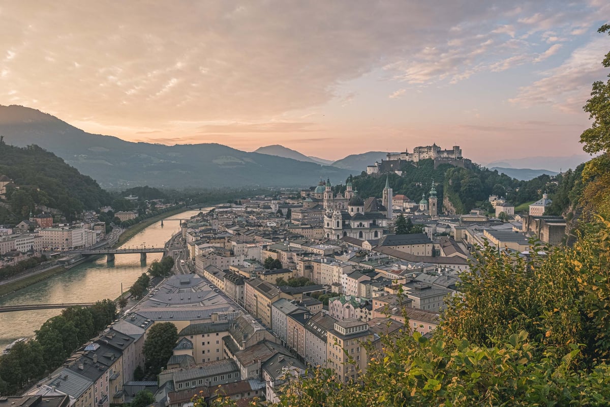 Panoramic sunrise view of Salzburg old town with cathedral domes and Hohensalzburg Fortress on snowy hills.