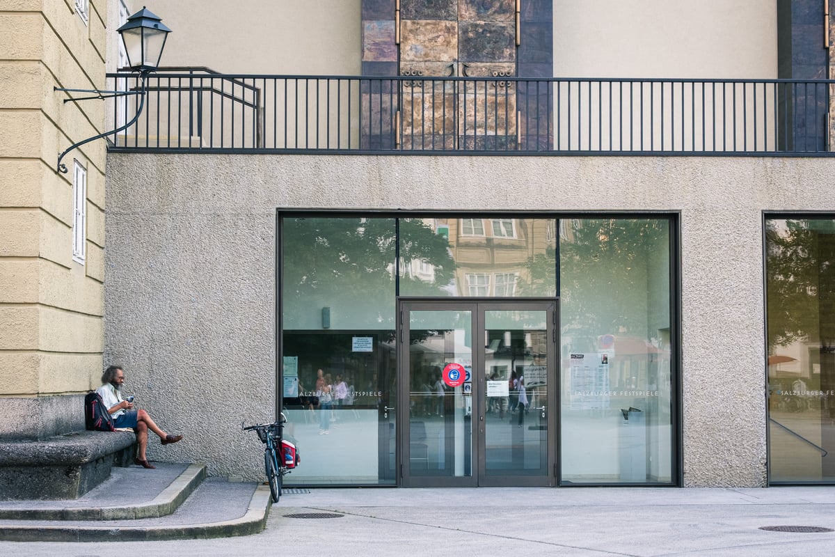 A man sits alone on a stone step beside a glass festival entrance lettered SALZBURGER FESTSPIELE, where a poster announces 100 Jahre Salzburger Festspiele, 1 to 30 August 2020.