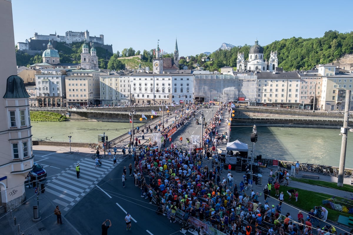 Thousands of runners are packed behind the start gantry and race clock at the head of the Staatsbruecke, with the Hohensalzburg Fortress and the Altstadt skyline across the Salzach.