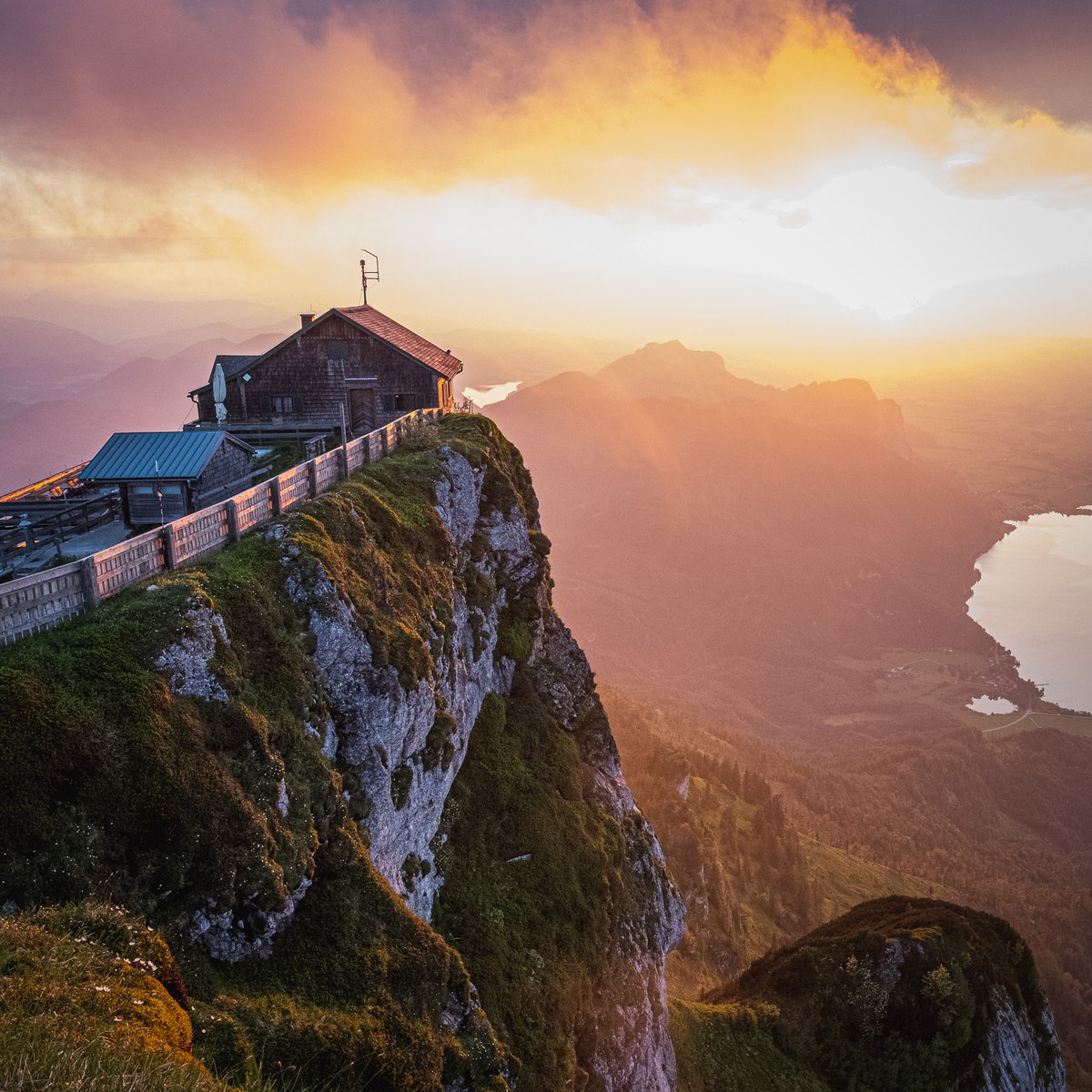 The shingled summit house stands right on the lip of the sheer Schafberg cliff, its stone-walled walkway running along the edge, with forested ridges and a lake far below in warm sunset light.