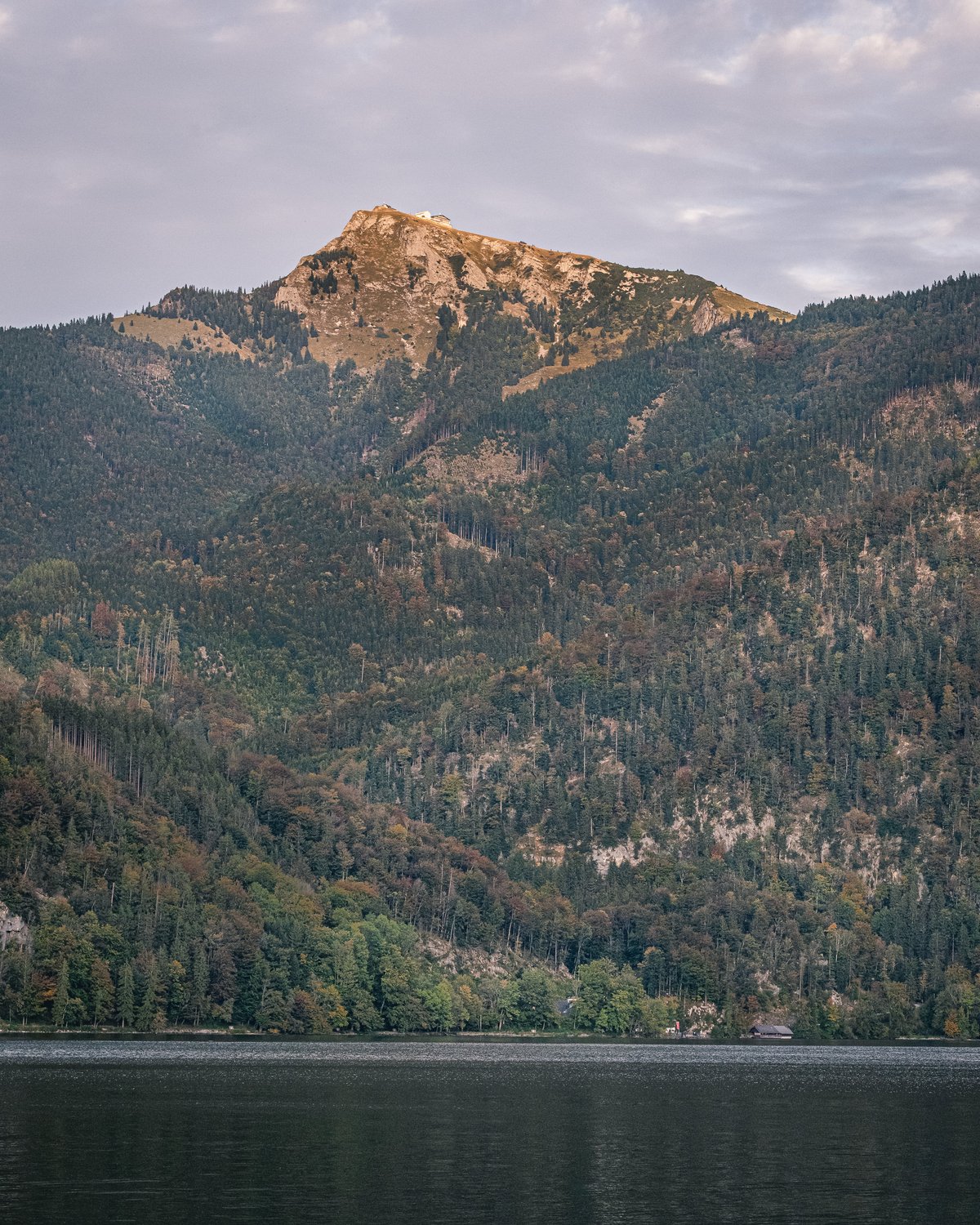The rocky Schafberg summit, with the white summit hotel on its crest, catches the last evening sun above a long forested flank that drops to the dark surface of Wolfgangsee.