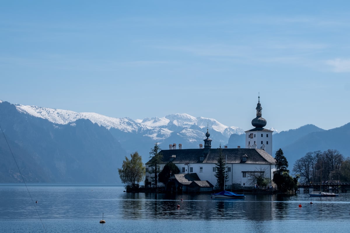 Seeschloss Ort sits on its wooded island in the Traunsee at Gmunden, its white onion-domed tower set against snow-covered peaks across the hazy blue lake.