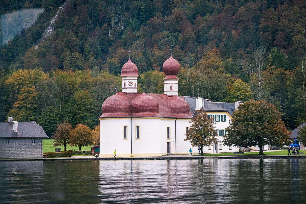 The red onion domes of the St. Bartholomae chapel rise from its white walls on the Koenigssee shore, backed by an autumn hillside and reflected in the lake.