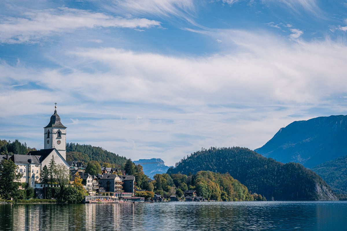 St. Wolfgang seen across the Wolfgangsee, the white pilgrimage church and its onion-domed tower rising above lakeside houses under a streaked blue sky.