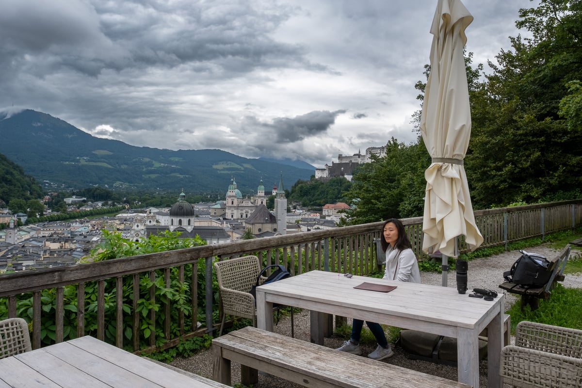 The gravel terrace of the Stadtalm on the Mönchsberg under heavy grey cloud, its weathered tables, benches and a furled umbrella looking out across the Salzburg Altstadt to Hohensalzburg on its ridge.